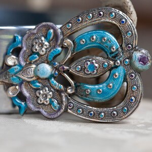 May include: A silver and blue enamel brooch with a floral design. The brooch is decorated with small, round, blue and purple stones.