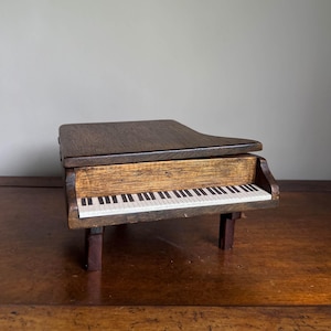May include: A miniature grand piano music box crafted from dark brown wood with a visible grain. The instrument features a flat top, a keyboard with alternating black and white keys, and stands on three sturdy wooden legs. The design is compact.