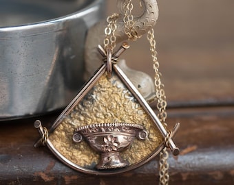 May include: A gold pendant necklace with a triangular frame and a small, ornate, gold cup charm. The pendant is hanging from a gold chain.