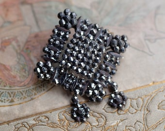 May include: A silver-toned brooch with a geometric design made of small, faceted beads. The brooch has a square shape with a pointed center and a dangling drop.