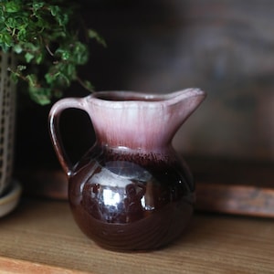 May include: A small ceramic pitcher featuring a rounded body with a glossy dark brown glaze on the lower half and a lighter, soft pinkish-white drip glaze effect around the rim and handle, complete with a curved spout and a side handle.