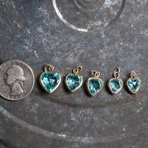 Puede incluir: Seis colgantes de coraz&oacute;n de oro con piedras preciosas azules de diferentes tama&ntilde;os. Los colgantes est&aacute;n dispuestos en una fila sobre una superficie de metal texturizada.