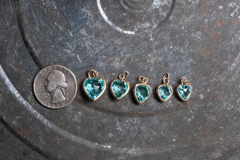 Puede incluir: Seis colgantes de coraz&oacute;n de oro con piedras preciosas azules de diferentes tama&ntilde;os. Los colgantes est&aacute;n dispuestos en una fila sobre una superficie de metal texturizada.