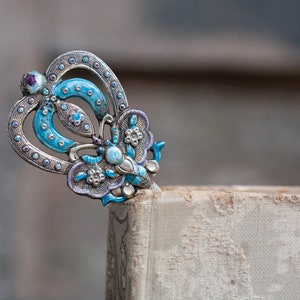 May include: A silver and blue butterfly-shaped bookmark with intricate details. The bookmark is made of metal and has a blue enamel finish. It is decorated with small white and purple gemstones.