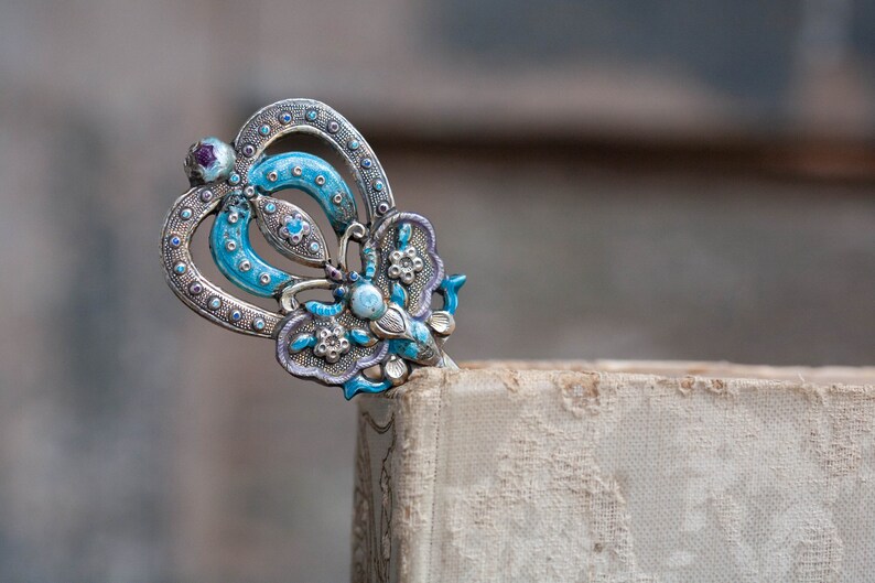 May include: A silver and blue butterfly-shaped bookmark with intricate details. The bookmark is made of metal and has a blue enamel finish. It is decorated with small white and purple gemstones.