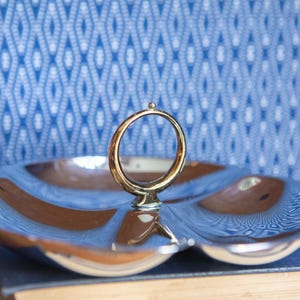 May include: A silver-toned metal serving dish with three distinct rounded compartments and a central circular gold-toned handle. The polished surface reflects light, creating a smooth, metallic appearance across the entire divided tray structure.