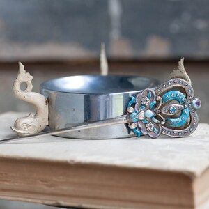 May include: A silver hair stick with a blue and white enamel design and a white ceramic dolphin head on the end. The stick is resting on a book.