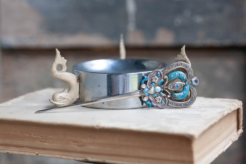 May include: A silver hair stick with a blue and white enamel design and a white ceramic dolphin head on the end. The stick is resting on a book.
