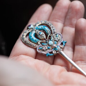 May include: A silver hair stick with a blue and white enamel design. The top of the stick is shaped like a butterfly with a crown on top.