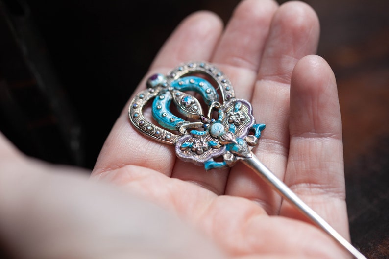 May include: A silver hair stick with a blue and white enamel design. The top of the stick is shaped like a butterfly with a crown on top.