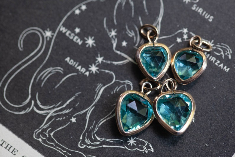 Puede incluir: Cuatro dijes en forma de coraz&oacute;n azul turquesa con engastes dorados. Los dijes est&aacute;n dispuestos en un grupo sobre un fondo negro con un dise&ntilde;o de constelaci&oacute;n de estrellas blancas.
