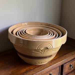 May include: A set of five nested mixing bowls made of glazed yellow ceramic. Each bowl features a wide rim and a decorative embossed arch pattern around the upper exterior, with a single horizontal brown stripe encircling the middle section.