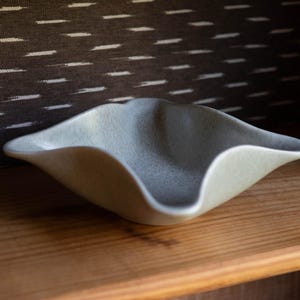 May include: A small, shallow ceramic bowl with a soft, matte, light gray finish. The bowl features a distinct, undulating, wavy rim that creates a four-cornered, organic shape. The interior surface has a subtle, speckled texture throughout.