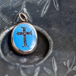 May include: An oval-shaped locket with a blue enamel background and a gold cross decorated with red gemstones. The locket is on a dark gray surface.