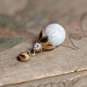 May include: A gold-toned pendant featuring a round, iridescent opal gemstone. The pendant has a floral design with three petals and a small, clear gemstone accent. The jewelry is displayed on a textured, neutral-toned surface.