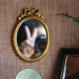 May include: An oval-shaped wall mirror with a metallic gold frame. The top of the frame features a decorative bow detail. The mirror surface has a beaded edge and reflects a hand making a peace sign and a camera lens in the center of the glass.