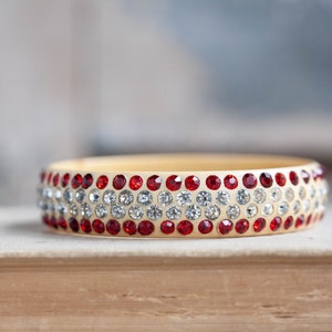 Puede incluir: Un brazalete dorado con piedras de imitación rojas y transparentes. El brazalete está sobre una superficie marrón claro.