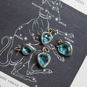 Puede incluir: Cuatro colgantes en forma de coraz&oacute;n con piedras preciosas facetadas de color azul claro. Los colgantes est&aacute;n sobre un fondo blanco con una ilustraci&oacute;n en blanco y negro de una constelaci&oacute;n.