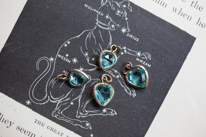 Puede incluir: Cuatro colgantes en forma de coraz&oacute;n con piedras preciosas facetadas de color azul claro. Los colgantes est&aacute;n sobre un fondo blanco con una ilustraci&oacute;n en blanco y negro de una constelaci&oacute;n.