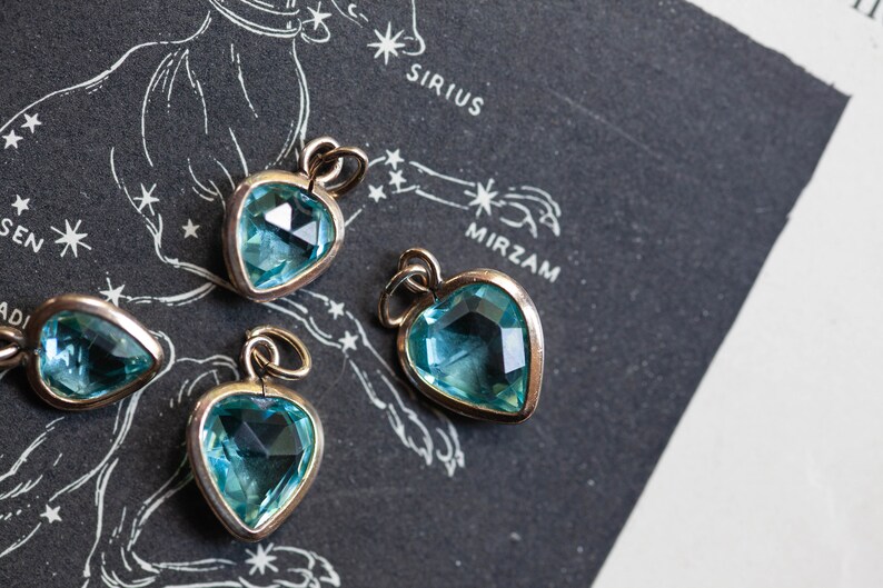Puede incluir: Cuatro colgantes en forma de coraz&oacute;n con detalles de piedras preciosas azules. Los colgantes est&aacute;n sobre un fondo negro con un mapa de estrellas impreso en &eacute;l. El mapa incluye los nombres Sirius y Mirzam.