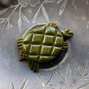 May include: A green plastic turtle brooch with a textured shell. The brooch is on a gray background with a floral design.