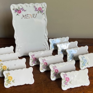 May include: A white ceramic menu board with scalloped edges and floral accents in pink, purple, and yellow, featuring the word MENU in gold. Accompanied by ten matching rectangular place card holders with similar floral patterns and scalloped edges.