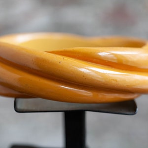 Puede incluir: Un brazalete de plástico amarillo con un diseño retorcido. El brazalete está en un soporte negro.