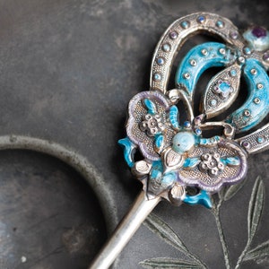 May include: A silver and blue hair ornament with intricate floral designs and a turquoise stone. The ornament is on a dark grey background.