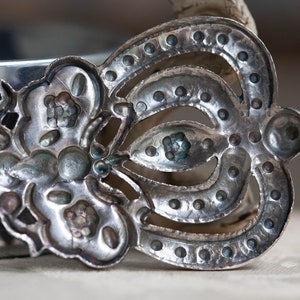 May include: A silver-toned metal spoon with a decorative design. The spoon has a floral pattern on one side and a crown-like design on the other side.