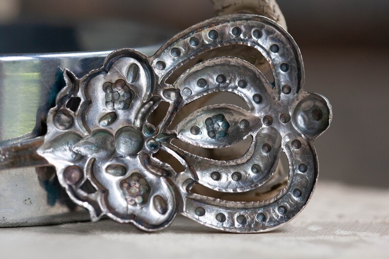 May include: A silver-toned metal spoon with a decorative design. The spoon has a floral pattern on one side and a crown-like design on the other side.
