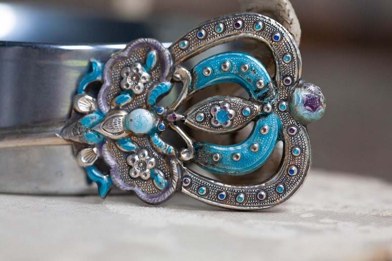 May include: A silver and blue enamel brooch with a floral design. The brooch is decorated with small, round, blue and purple stones.