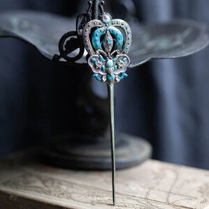 May include: A silver hair stick featuring a blue and white enamel butterfly design. The butterfly has a crown on its head and is surrounded by small, clear rhinestones.