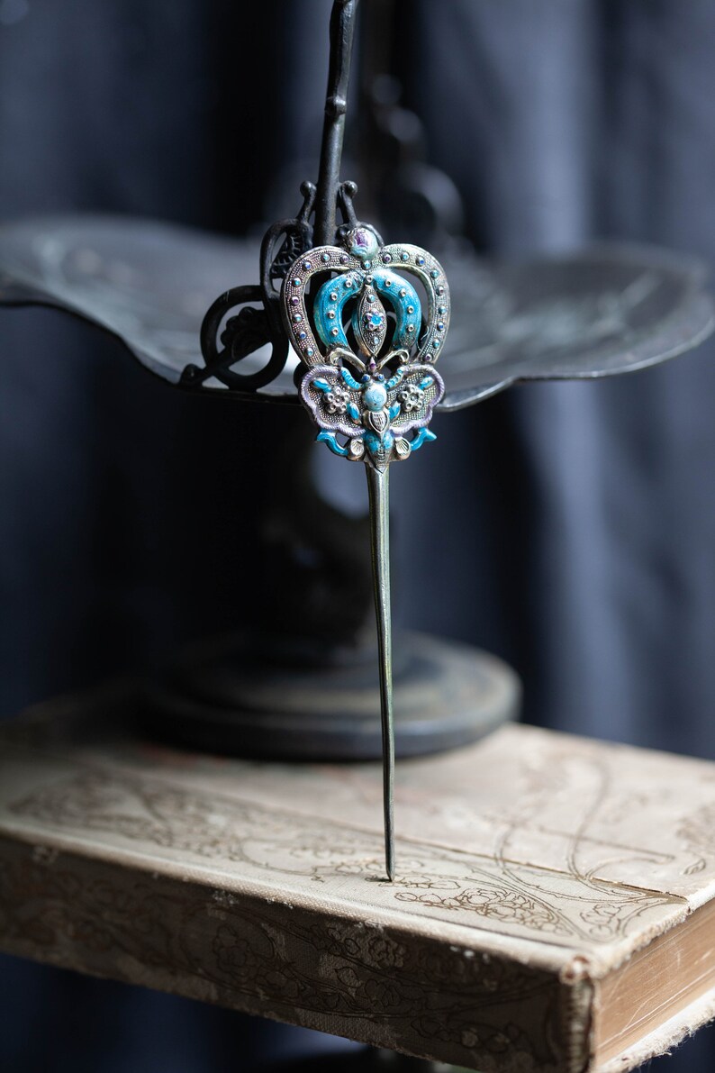 May include: A silver hair stick featuring a blue and white enamel butterfly design. The butterfly has a crown on its head and is surrounded by small, clear rhinestones.