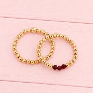 May include: Two circular beaded rings constructed from small, polished gold-toned spherical beads. One ring consists entirely of gold beads, while the second ring features a series of gold beads accented by three small, faceted dark red beads.