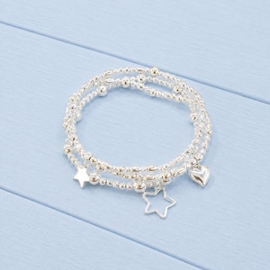 May include: A multi-strand silver-toned beaded bracelet featuring small round metallic beads. The bracelet includes three dangling charms: a solid five-pointed star, an open-frame five-pointed star, and a smooth, rounded heart-shaped charm.