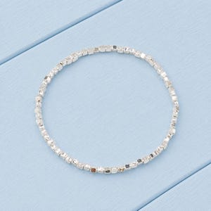 May include: A circular bracelet composed of numerous small, metallic silver-toned cube-shaped beads strung together. The beads have a reflective, polished finish and are arranged in a continuous, uniform loop, creating a simple and sleek jewellery piece.