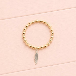 May include: A circular stretch bracelet composed of small, polished gold-toned spherical beads. A single silver-toned metal charm shaped like a detailed feather hangs from the bottom of the bracelet, centred at the base of the circular band.