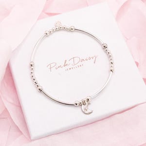 May include: A silver bracelet featuring a crescent moon charm and small beads. The bracelet rests on a white box with the text "Pink Daisy Jewellery" in pink script. The background is a soft pink fabric.