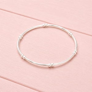 May include: A circular silver bracelet featuring a series of small, smooth metallic beads spaced evenly along the thin, polished band. The design consists of alternating segments of plain wire and pairs of tiny spherical accents in a continuous loop.