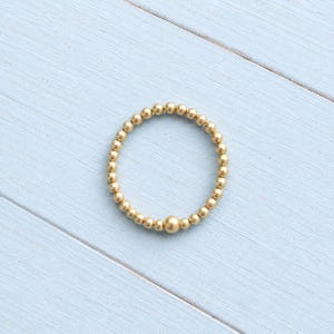 May include: A circular ring composed of small, uniform gold-toned spherical beads arranged in a continuous loop, featuring one slightly larger gold-toned bead positioned at the bottom centre of the circular band design.