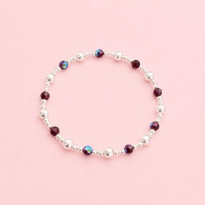 Puede incluir: Una pulsera de cuentas circular que presenta un patrón repetitivo de pequeñas esferas metálicas plateadas pulidas intercaladas con cuentas más grandes, facetadas y de color burdeos oscuro con sutiles reflejos iridiscentes azules y morados.