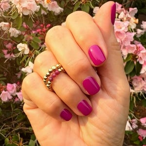 Può includere: Una mano con unghie corte color magenta e un anello d'oro con piccole perline rotonde. L'anello ha una fila di perline dorate e una fila di perline rosse. Fiori rosa sullo sfondo.