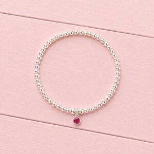May include: A circular stretch bracelet composed of small, polished silver-toned beads. A single, round, vibrant pink gemstone charm is suspended from the bottom of the bracelet, featuring a simple silver-toned bezel setting that secures the stone.
