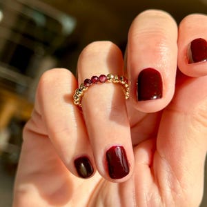 May include: A gold ring with small red beads. The ring is on a finger with dark red nail polish.