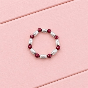 May include: A circular ring featuring an alternating pattern of small, faceted, deep red spherical beads and smooth, elongated, metallic silver-toned cylindrical beads arranged in a continuous loop on a flexible cord.