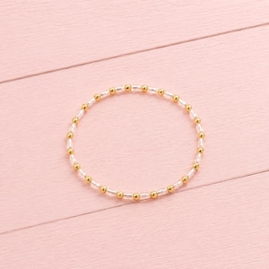 May include: A circular beaded bracelet featuring an alternating pattern of small, reflective gold-toned metallic spheres and clear, translucent glass beads arranged in a continuous loop to form a simple, delicate piece of jewellery.