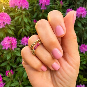 Peut inclure: Une bague composée d'un anneau de petites perles dorées polies, agrémenté de trois minuscules perles de pierre précieuse facettées rose foncé. La bague est portée sur un doigt aux ongles vernis de rose clair, devant des fleurs de rhododendron.