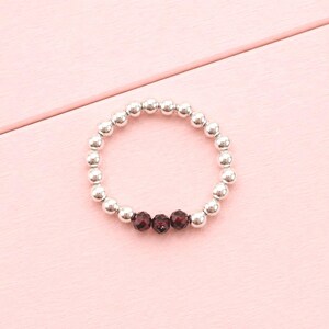May include: A circular stretch ring composed of numerous small, polished silver-toned metallic beads. The ring features three dark red, faceted, round beads positioned together at the bottom centre of the circular band design.
