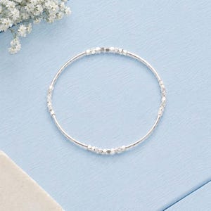 May include: A circular silver bracelet featuring a series of small, polished metallic cube-shaped beads spaced evenly along a thin, flexible wire band. The design is simple and uniform, showcasing a reflective finish on each individual cube component.
