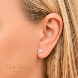 Puede incluir: Primer plano de una oreja con un pequeño pendiente de cristal transparente, redondo. El pendiente está engarzado en metal plateado o blanco. El texto "Pink Daisy Jewellery" es visible en la esquina inferior izquierda.